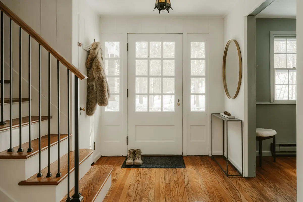 Recibidor luminoso con escalera de madera, abrigo colgado y puerta de cristal